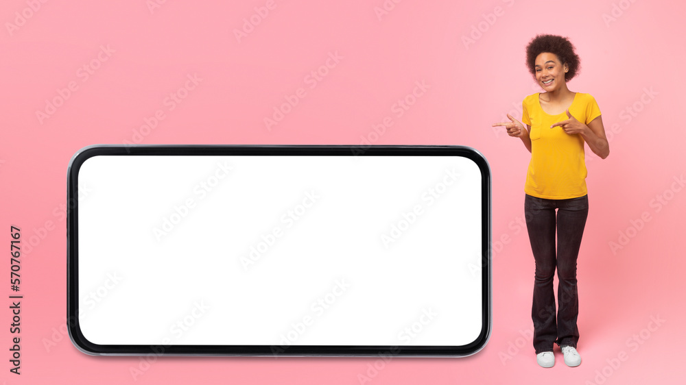Glad young african american curly female in casual points fingers at huge smartphone with blank screen Glad young african american curly female in casual points fingers at huge smartphone with blank screen