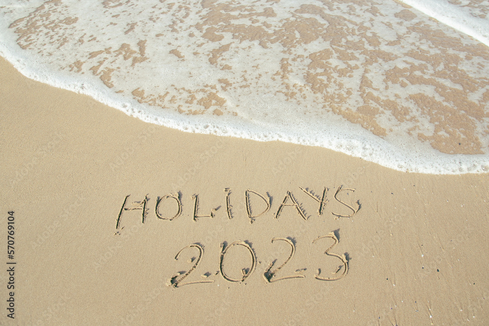 Holidays 2023 lettering on the beach with wave and clear blue sea. 