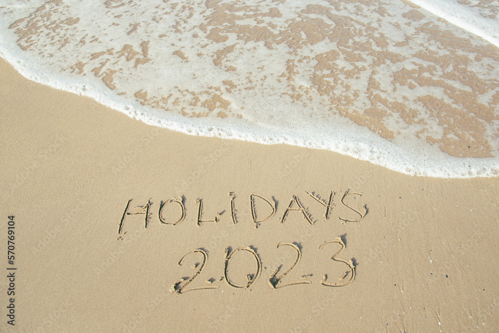 Holidays 2023 lettering on the beach with wave and clear blue sea. Holidays 2023 lettering on the beach with wave and clear blue sea.