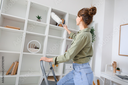 Young woman on stepladder cleaning bookshelf with duster at home