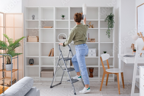 Young woman on stepladder cleaning bookshelf with duster at home
