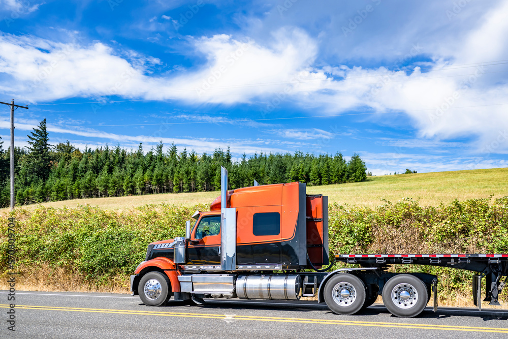 Stylish black and orange big rig semi truck transporting empty flat bed semi trailer driving on the road along the summer meadow with green grass and line of trees