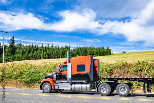 Stylish black and orange big rig semi truck transporting empty flat bed semi trailer driving on the road along the summer meadow with green grass and line of trees