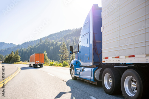 Two big rigs semi trucks with refrigerator and dry van semi trailers driving in convoy on the winding highway road at sunlight