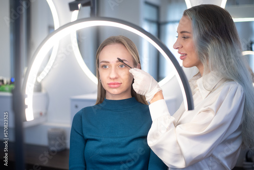 Make up artist applying make up. Young woman in cosmetology salon receive face care consultation. Beautician shows how care for eyebrows. Every woman wants look attractive at any age.