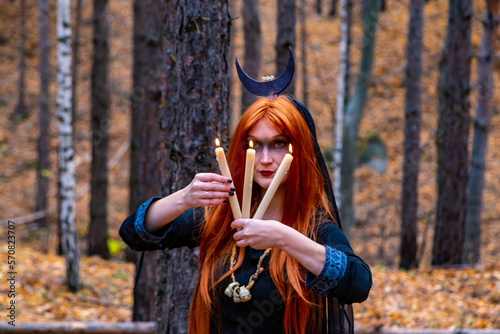 Gothic image of a red-haired girl on a fashion shoot in the forest in a black dress with a tiara on her head and three candles burning in the background pine forest