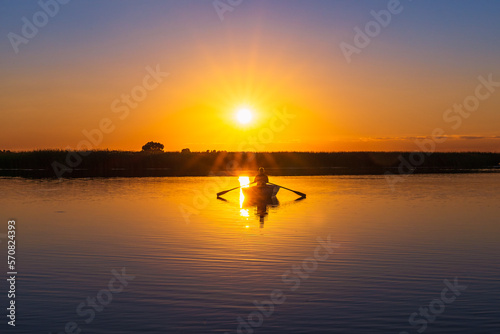A classic rowboat against a backlit orange sunset in semi-darkness. Silhouette of a 60-year-old Caucasian male with oars in his hands in the rays of the sun