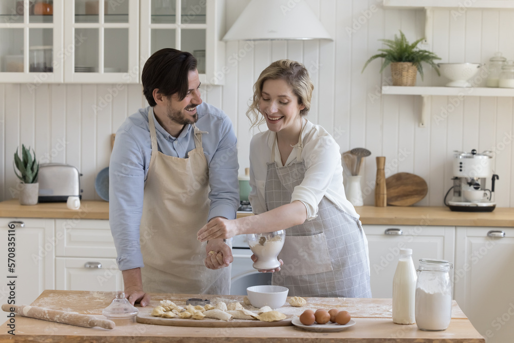 Happy positive young couple in love wearing aprons, enjoying bakery food preparation, baking pies in home kitchen, sifting flour on dough at table with ingredients, talking, smiling