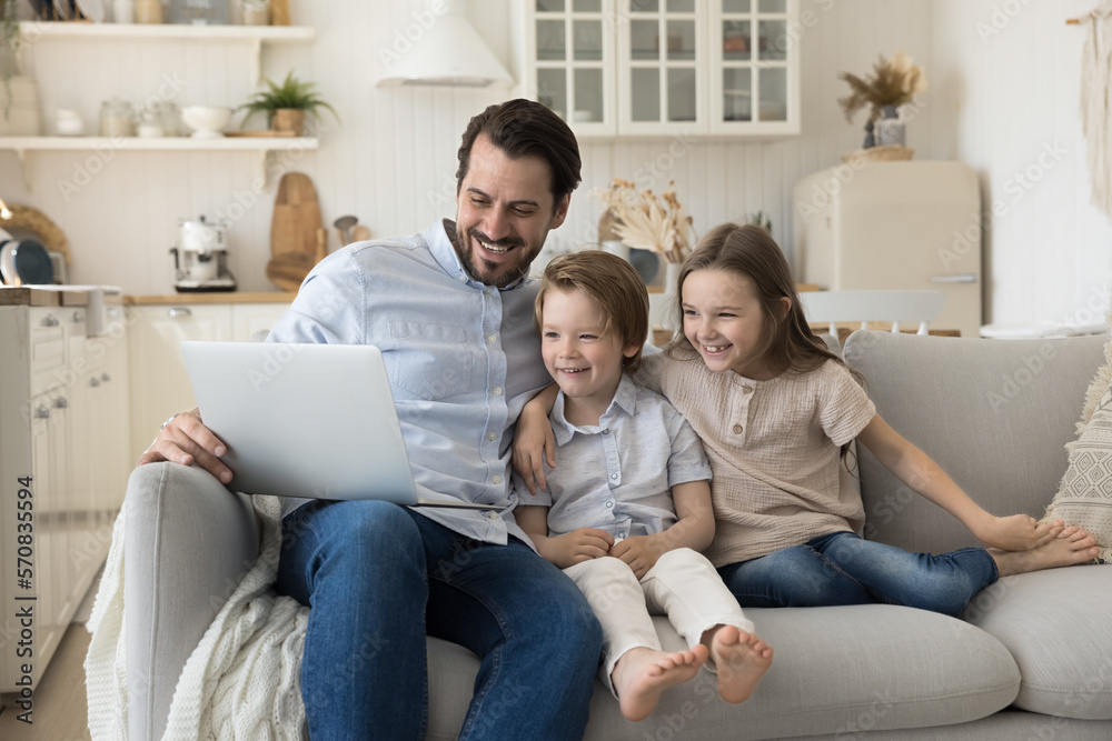 Happy father and two joyful little kids using media app on laptop for Internet communication, watching cartoon funny movie, online TV family channel, resting on sofa at home
