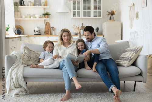 Positive engaged couple of parents teaching two cute kids to read book, watching pictures, telling funny fairytale, relaxing on home couch, hugging children, enjoying literature hobby