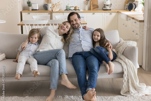 Happy mom, dad and two little kids resting on soft couch together, hugging with love, care, looking at camera away, smiling, laughing. Parents embracing children. Home portrait