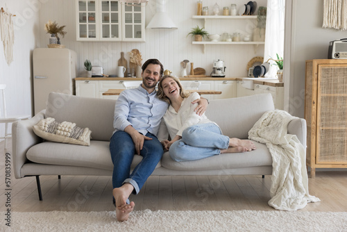 Happy young married couple in love enjoying being at new cozy home, celebrating real estate property owning, resting on comfortable couch in modern living room. Wide shot portrait photo