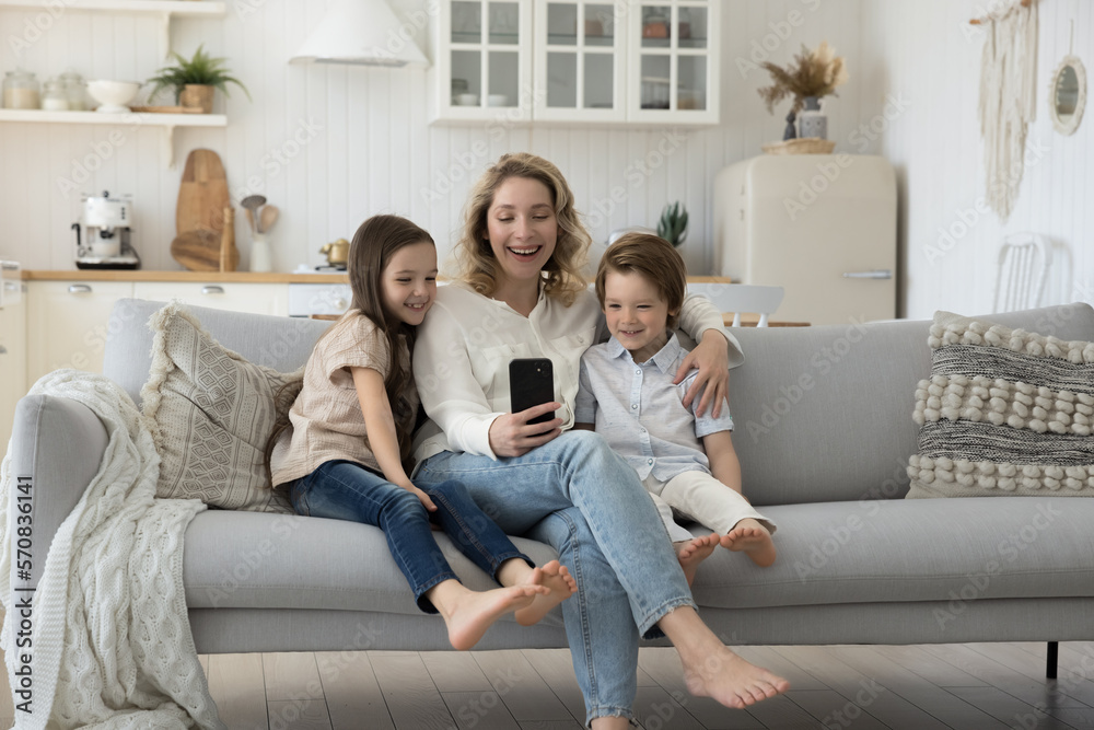 Positive blogger mom hugging two little sibling children on home sofa, using mobile phone for Internet communication, talking on video call, taking family selfie picture Positive blogger mom hugging two little sibling children on home sofa, using mobile phone for Internet communication, talking on video call, taking family selfie picture