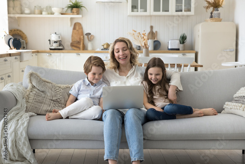 Cheerful young daycare sitter entertaining children at home, watching educational cartoon on laptop. Happy mother hugging little kids on sofa, enjoying funny movie, online communication