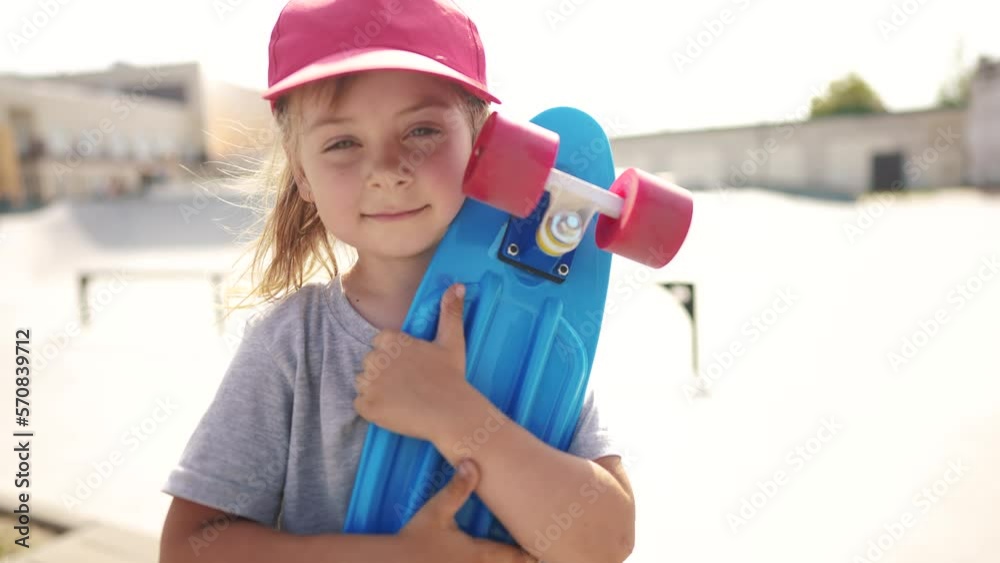child with a skateboard. girl in a red cap with a skateboard on the playground portrait. skateboarder child close-up outdoors sun glare. kid skateboarder looking lifestyle at the camera child with a skateboard. girl in a red cap with a skateboard on the playground portrait. skateboarder child close-up outdoors sun glare. kid skateboarder looking lifestyle at the camera