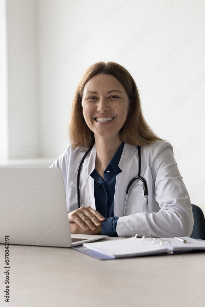 Happy pretty doctor woman sitting at work table in hospital, clinic office, looking at camera with toothy smile. Portrait of positive cheerful female practitioner, physician in white coat, stethoscope Happy pretty doctor woman sitting at work table in hospital, clinic office, looking at camera with toothy smile. Portrait of positive cheerful female practitioner, physician in white coat, stethoscope