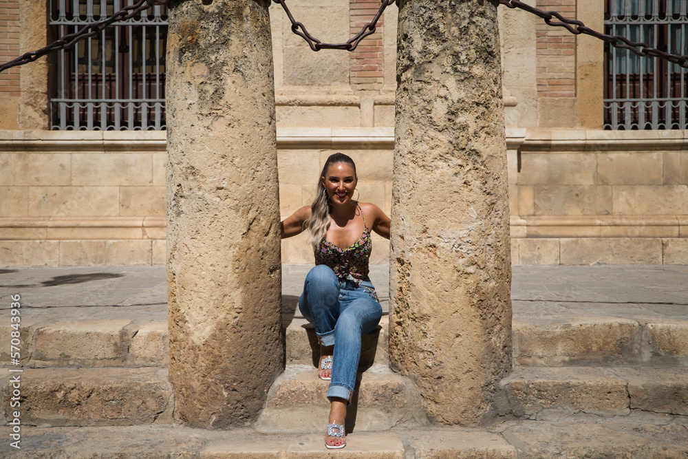 Beautiful young blonde Spanish woman is sitting between two columns of the cathedral of Seville. The woman is dressed in modern clothes and is happy.