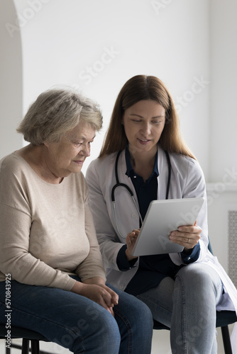 Friendly positive GP doctor showing tablet screen to elderly 80s patient woman, explaining examination result, treatment, roentgen electronic shot, giving consultation, expertise on appointment