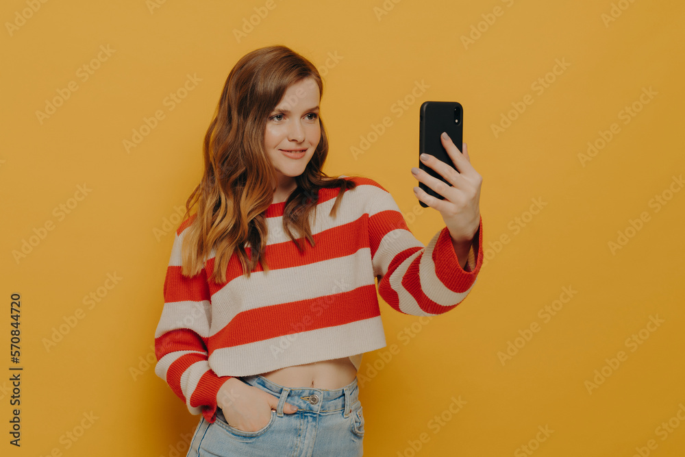 Naklejka premium Young charming woman blogger talking selfie on mobile phone while posing on yellow background