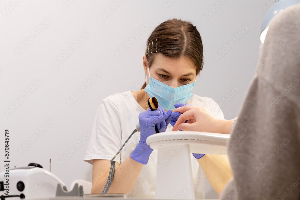 White nail technician in mask drills, removes client's cuticle with hardware machine, holding nail brush. Customer wears warm cloth, sweater. Cold season. Combined manicure, beauty treatment in spa