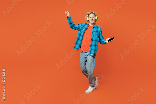 Full body young blond fun caucasian man wear blue shirt orange t-shirt headphones listen to music use mobile cell phone dance isolated on plain red background studio portrait People lifestyle concept