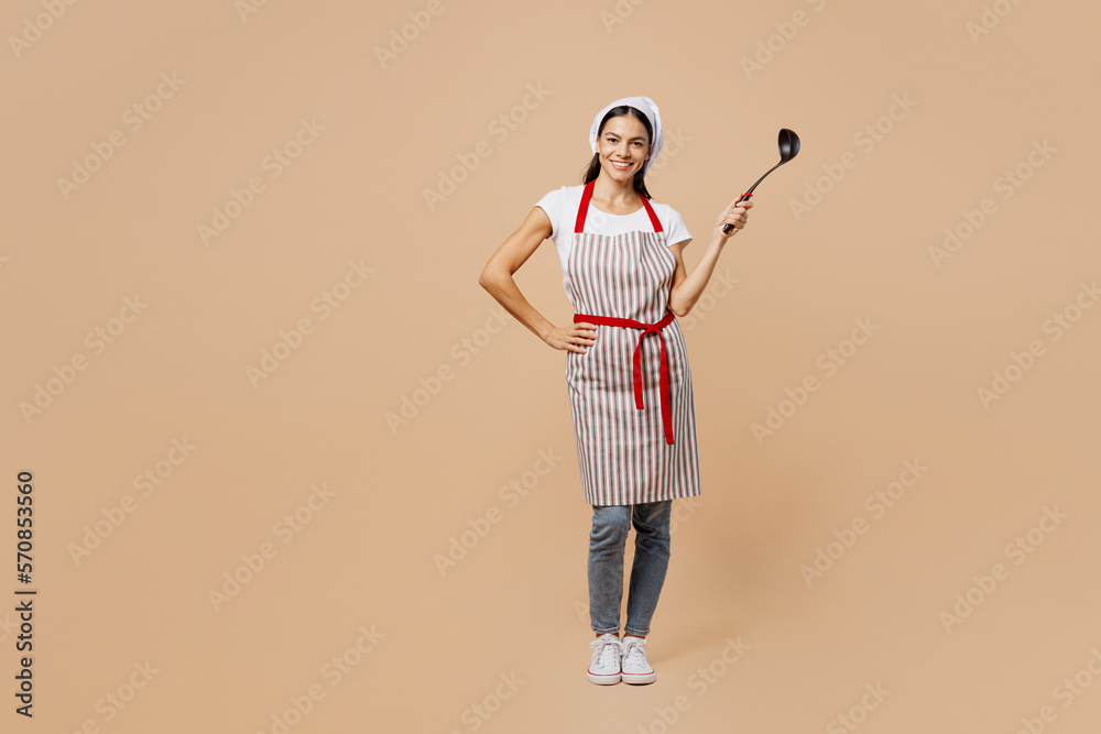 Full body smilng happy fun young housewife housekeeper chef baker latin woman wear apron toque hat hold in hand laddle look camera isolated on plain pastel light beige background. Cook food concept. Full body smilng happy fun young housewife housekeeper chef baker latin woman wear apron toque hat hold in hand laddle look camera isolated on plain pastel light beige background. Cook food concept.
