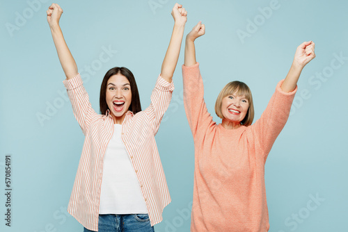 Lucky happy elder parent mom with young adult daughter two women together wear casual clothes do winner gesture clench fist raise up hands isolated on plain blue cyan background. Family day concept.