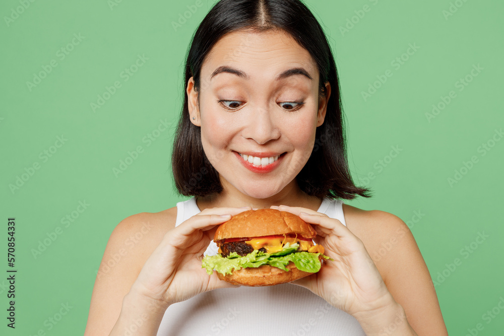 Close up young happy excited woman wearing white clothes holding eating biting tasty burger isolated on plain pastel light green background Proper nutrition healthy fast food unhealthy choice concept Close up young happy excited woman wearing white clothes holding eating biting tasty burger isolated on plain pastel light green background Proper nutrition healthy fast food unhealthy choice concept