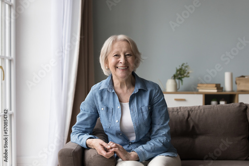 Fotografie Joyful positive senior lady sitting on sofa, smiling, laughing, looking at camera, enjoying retirement at cozy home