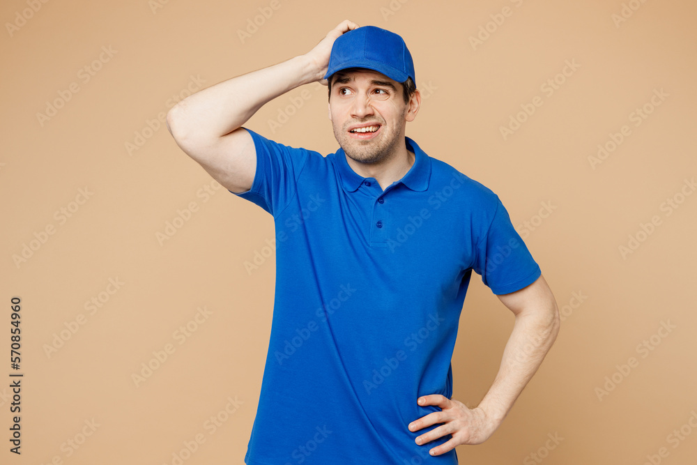 Professional mistaken sad delivery guy employee man wear blue cap t-shirt uniform workwear work as dealer courier hold scratch head look aside isolated on plain light beige background Service concept