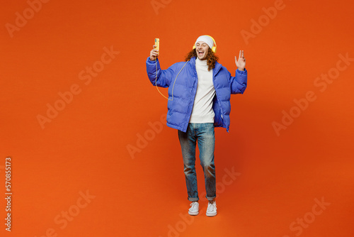 Full body young happy man with long curly hair wearing hat purple ski padded jacket casual clothes headphones listen to music use mobile phone isolated on plain orange red background studio portrait.