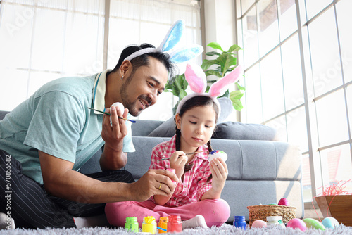Father and daughter preparing beautiful Easter eggs for decoration spring celebration, pink little bunny girl kid and dad with rabbit ears headband painting colorful eggs together, happy family Easter
