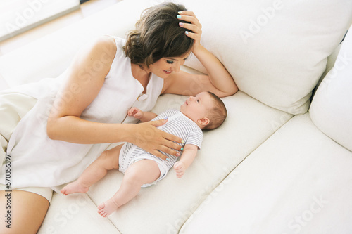 Indoor portrait of happy young mother playing with adorable little baby, lying on white couch