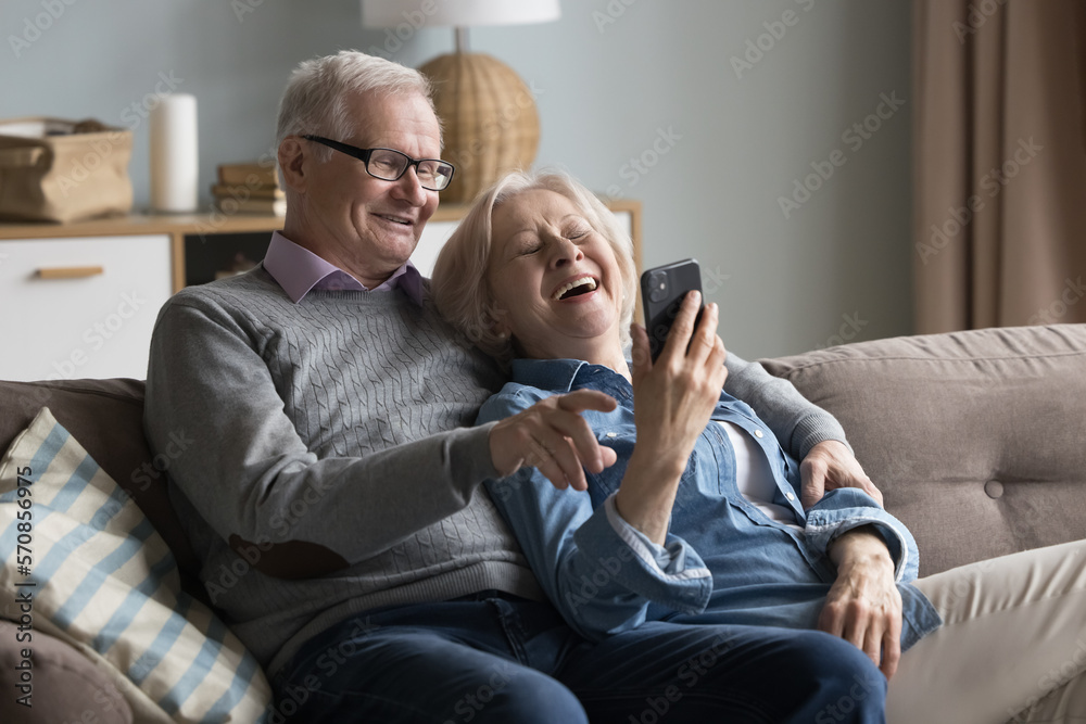 Joyful old husband and wife watching media on mobile phone, talking on video call together, smiling, laughing. Mature retired couple using online app for shopping, internet communication - obrazy, fototapety, plakaty 