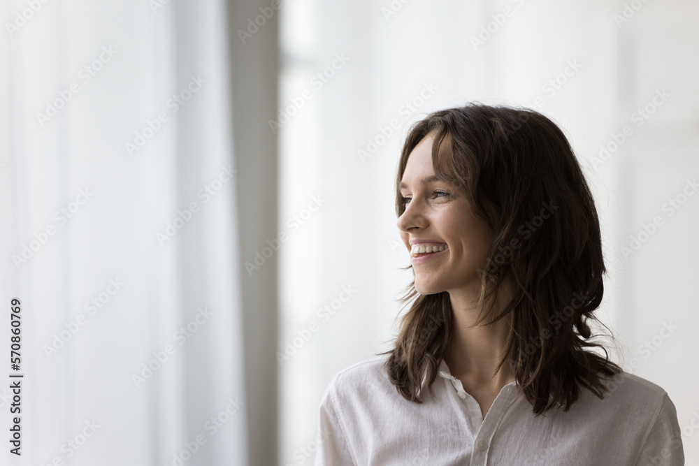 Happy dreamy young woman indoor candid portrait. Beautiful adult girl with healthy white teeth smiling, looking at window away, dreaming, thinking on good news, success