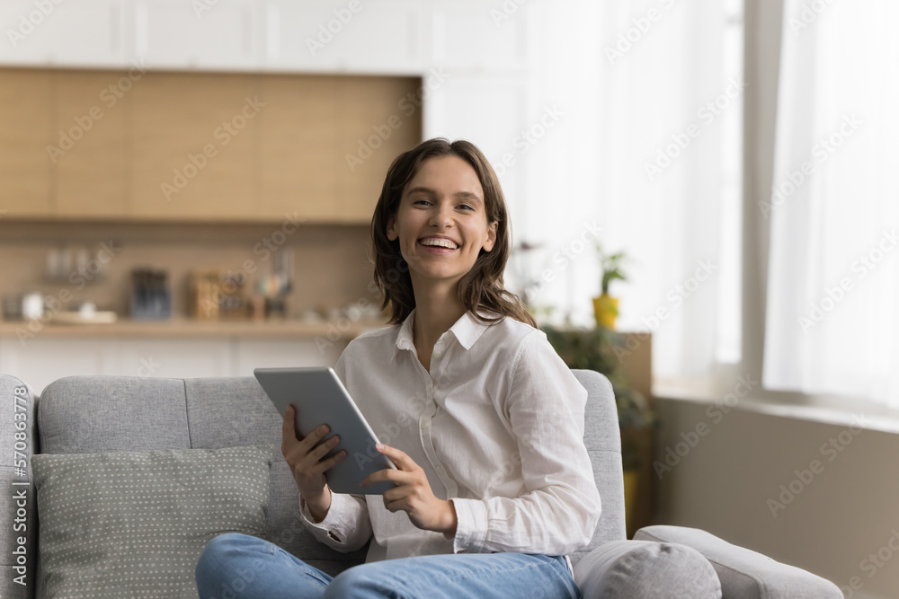 Happy pretty young tablet user woman home portrait. Beautiful young millennial blogger sitting on sofa, holding digital device, looking at camera with toothy smile, enjoying online communication Happy pretty young tablet user woman home portrait. Beautiful young millennial blogger sitting on sofa, holding digital device, looking at camera with toothy smile, enjoying online communication
