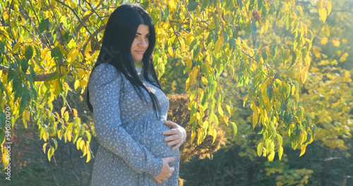 Happy pregnant woman smiling dreaming about baby in sunny autumn park. Pregnant mother strokes, touches belly on nature background of yellow trees foliage. Bright natural sun light day in fall forest