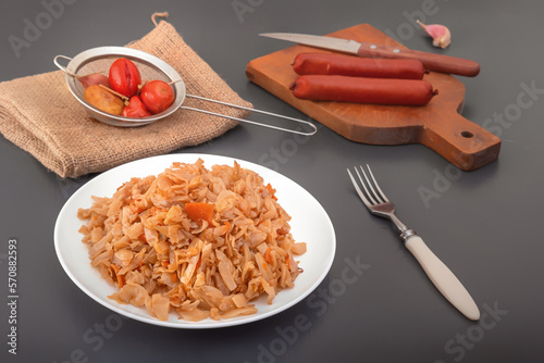 Stewed cabbage - bigos in a white porcelain plate against the background of smoked sausages and pickled vegetables