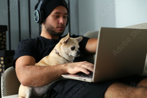 A young, stylish guy in black clothes is at home with his dog. A Chihuahua dog and a guy watch a movie together on a laptop, have fun and spend time together, real friends. The guy and the dog are in 