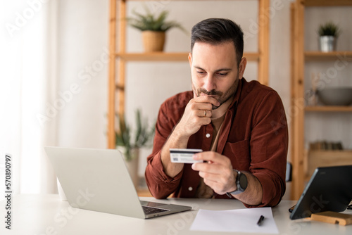 Serious business man sitting at table in home office, holding and looking at credit card, worried about debt and credit card deficit, considering a financial loan from bank. photo