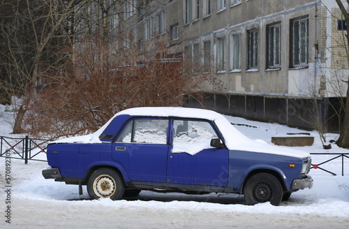 An old snow-covered blue Soviet car is parked on the street, Bolshevikov Avenue, St. Petersburg, Russia, february 2023