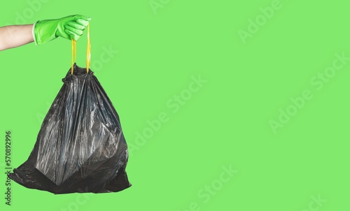 Hand in glove holding handles of black plastic disposable trash bag full of garbage, waste on green banner background with copy space for text photo