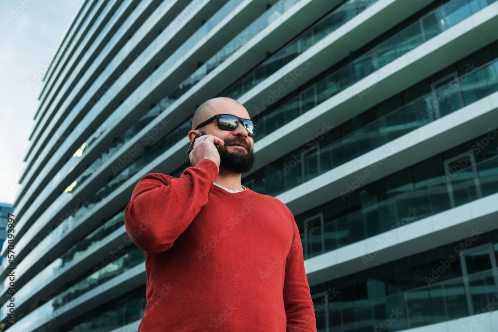Adult hipster bearded man in sunglasses talking on mobile phone outdoor the modern office buildings. IT guy Adult hipster bearded man in sunglasses talking on mobile phone outdoor the modern office buildings. IT guy