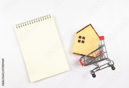 flat layout of wooden house model in shopping trolley or shopping cart with blank page opened notebook on white background, home purchase concept.