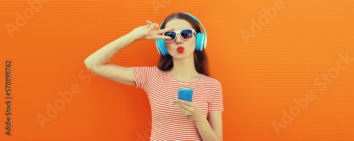 Portrait of stylish young woman in headphones listening to music with smartphone on orange background