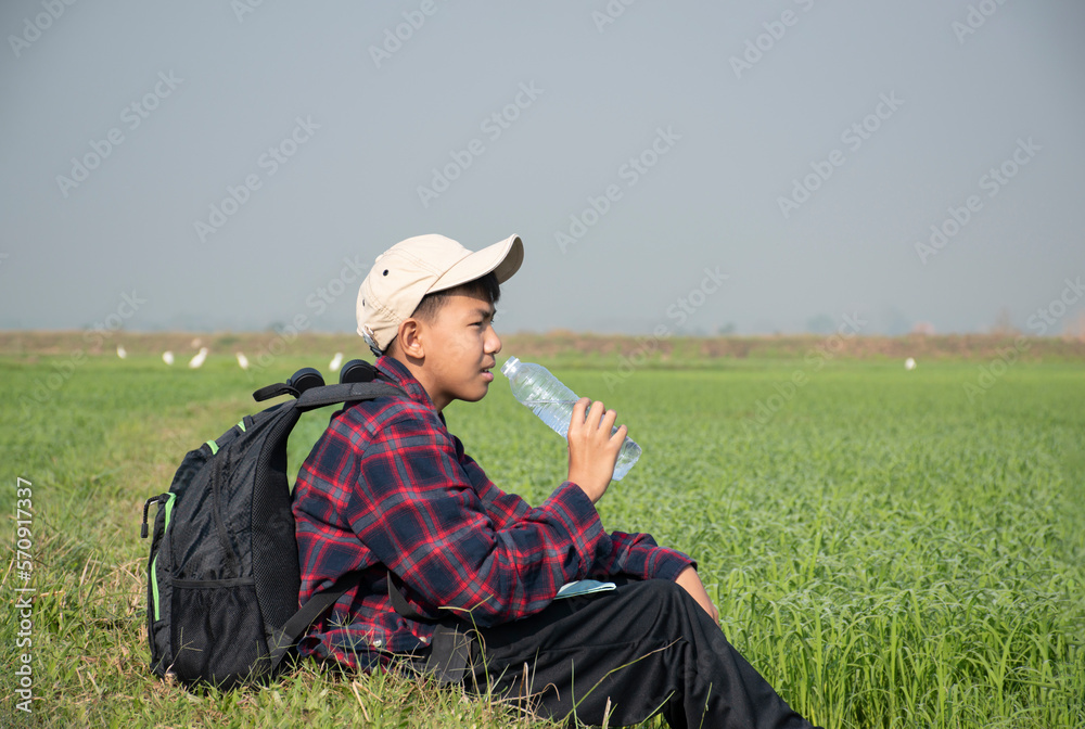 Asian boy in plaid shirt and has backpack wears cap and holds drinking water bottle, sitting on ridge rice field which has egrets behind during his birdwatching and his summer vacation. Asian boy in plaid shirt and has backpack wears cap and holds drinking water bottle, sitting on ridge rice field which has egrets behind during his birdwatching and his summer vacation.