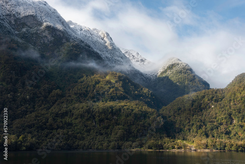 The diverse shores in the Doubtful Sound with spotlights of light, wispy clouds, and snowcapped rainforest.