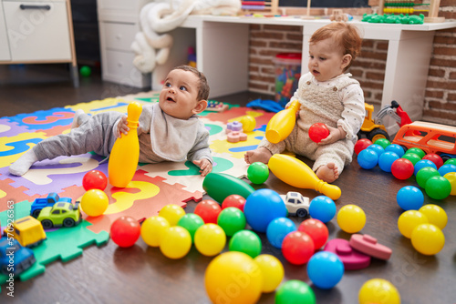 Two adorable babies playing with balls and bowling pin sitting on floor at kindergarten