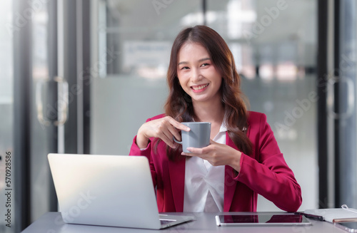 Pretty asian businesswoman sitting happily working on laptop and drinking coffee with bright smile in office.