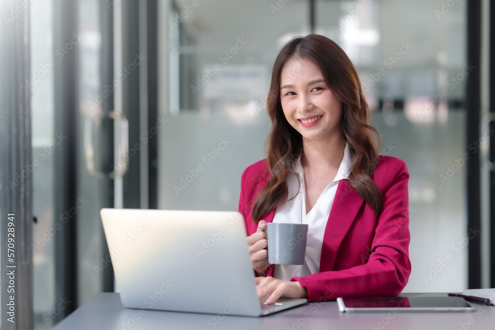 Pretty asian businesswoman sitting happily working on laptop and drinking coffee with bright smile in office. Pretty asian businesswoman sitting happily working on laptop and drinking coffee with bright smile in office.